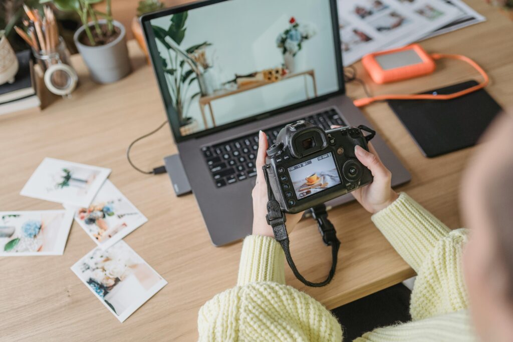 woman looking at the viewfinder of her camera as she sits in front of her laptop with photos laying on the desk.